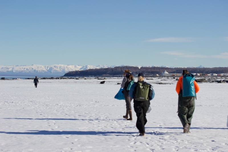 Students wearing dry bag backpacks walk across a snowy expanse with blue skies above and snow mountain peaks in the distance.